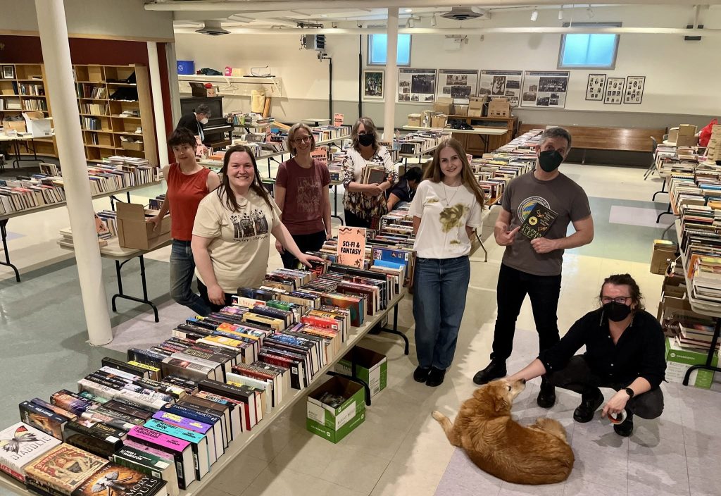 Seven unidentified volunteers are standing around table full of books. A dog is laying on the floor.